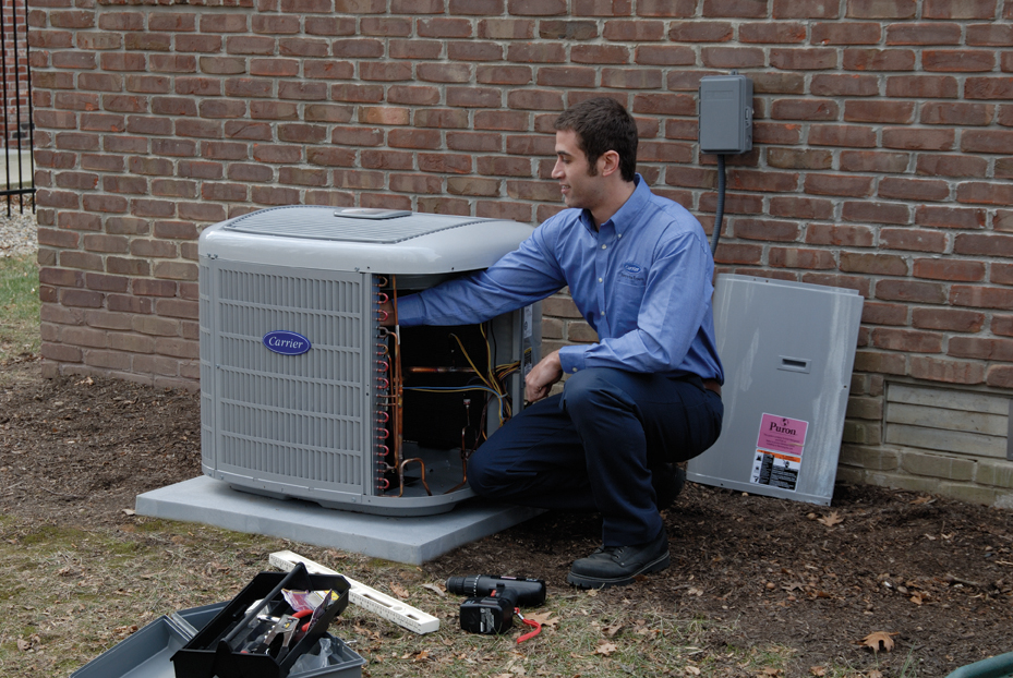 A technician in a blue shirt is kneeling beside an outdoor HVAC unit near a brick wall, ensuring the fairest deal for quality service, with tools scattered on the ground.