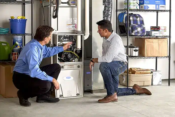 Two men kneel in a garage, examining a furnace or heating unit, surrounded by shelves with various stored items.