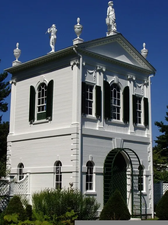A white two-story building in Danvers, adorned with green shutters and classical statues on the roof, stands elegantly against a clear blue sky.